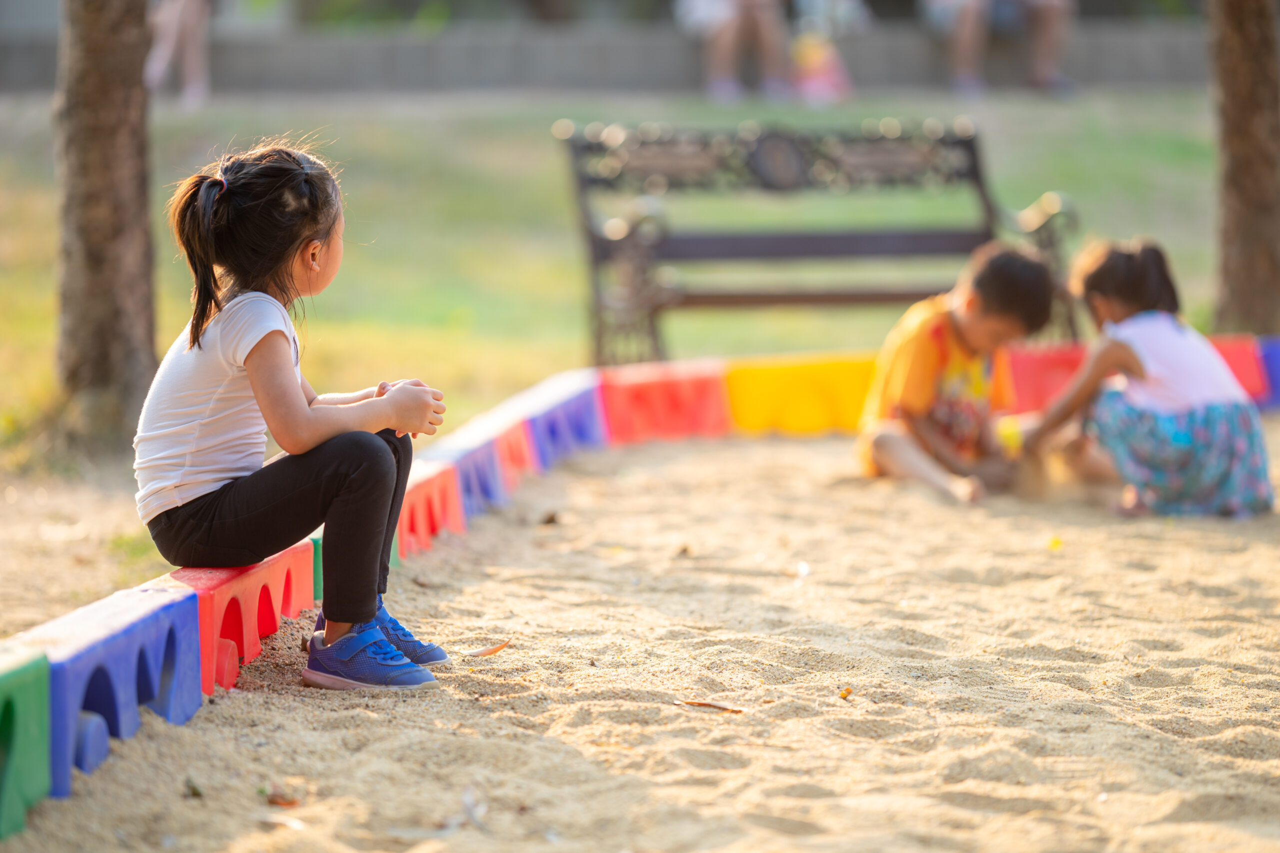 Little girl sitting lonely watching friends play at the playground