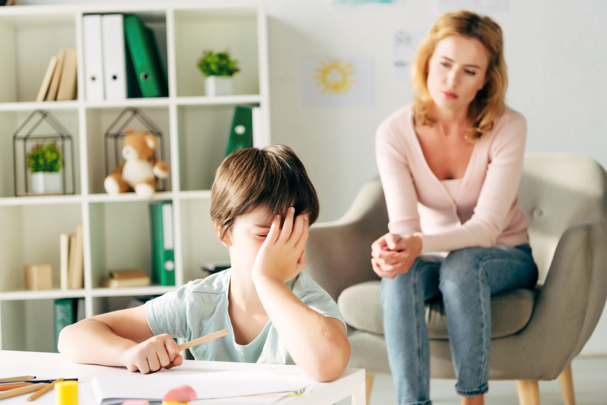 sad kid with dyslexia holding pencil and child psychologist on background